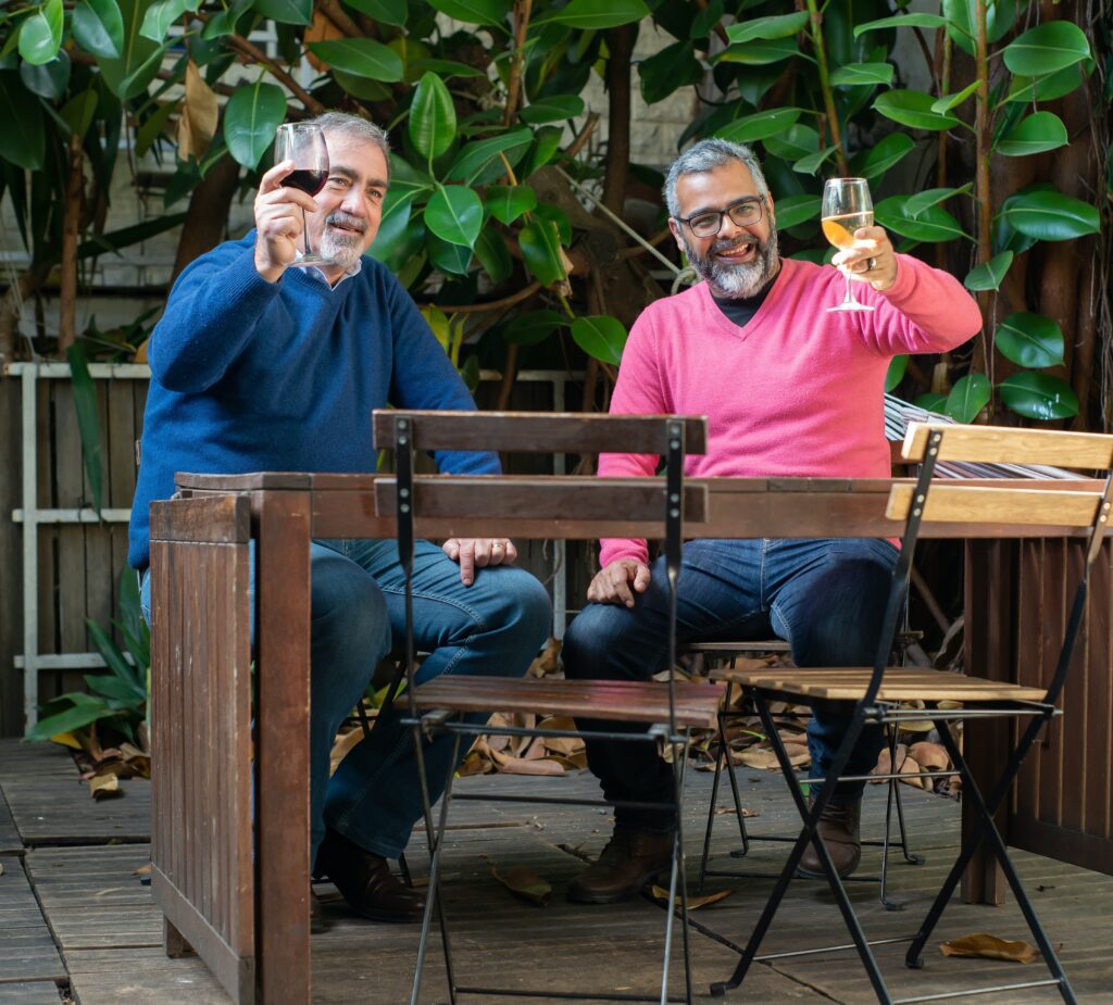 two adult men in 50s on porch with glasses of wine toasting in air 